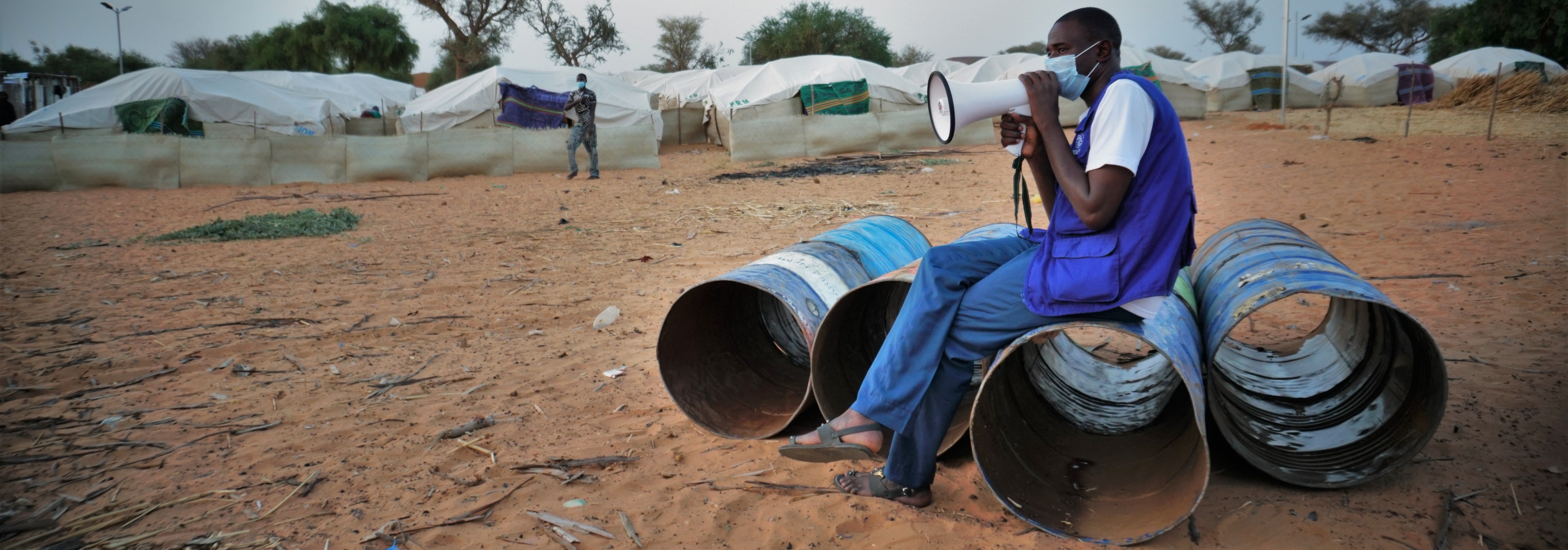 An IOM staff member wearing a protective mask is sitting on large empty metal cans and speaking into a megaphone. 