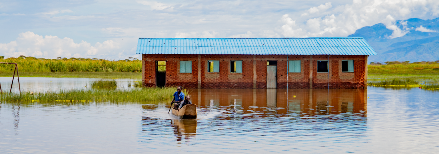Two men paddle through a schoolyard, past abandoned classrooms. Photo: ©IOM 2021/Triffin Ntore